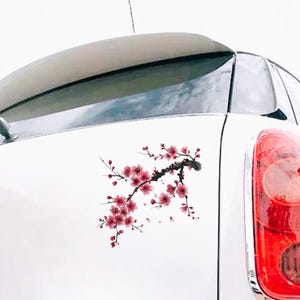May include: A white car with a cherry blossom branch decal. The decal features pink flowers and a dark brown branch. The car's taillight is visible on the right side of the image.