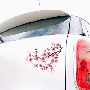 May include: A white car with a cherry blossom decal on the rear. The decal features pink flowers and brown branches, creating a heart shape. The car's taillight is visible on the right.