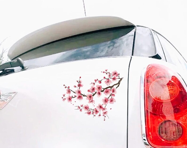May include: A white car with a cherry blossom decal on the rear. The decal features pink flowers and brown branches, creating a heart shape. The car's taillight is visible on the right.