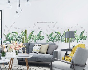 May include: A gray sofa with green and yellow throw pillows sits in front of a white wall with a green and white tropical plant wall decal. The decal features palm leaves, cacti, and dragonflies. The text "At the touch of love, everyone becomes a poet" is written on the wall.
