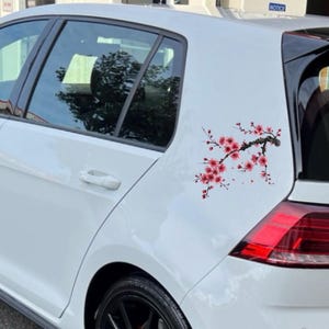 May include: A white car with a cherry blossom branch decal on the side. The decal features a dark brown branch with pink and red flowers. The car has black tires and tinted windows.