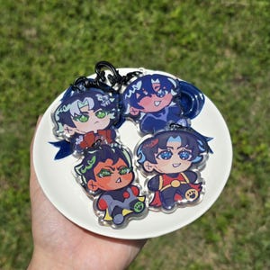 Könnte beinhalten: Vier Schlüsselanhänger mit Anime-Charakteren und schwarzen Schlüsselringen. Jeder Schlüsselanhänger zeigt ein anderes Charakterdesign mit leuchtenden Farben, darunter Blau, Rot, Grün und Gelb. Die Schlüsselanhänger sind auf einem weißen Teller mit blauen Akzenten angeordnet.