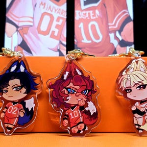 May include: Three acrylic keychains featuring anime-style characters with fox ears and tails. Each character wears a red and white sports uniform with a number. The keychains have gold-colored clasps and are set against an orange background.