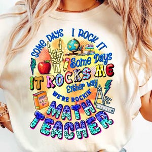May include: A cream-coloured t-shirt featuring a vibrant graphic. The design includes the text "Some Days I Rock It, Some Days It Rocks Me, Either Way We're Rockin' Math Teacher" with illustrations of an apple, a skeleton hand, a globe, and math tools.