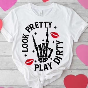 May include: White t-shirt featuring a black skeleton hand in a rock and roll pose. The text "LOOK PRETTY PLAY DIRTY" is printed around the hand, with red lipstick kiss marks.