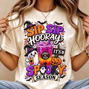 Puede incluir: Una camiseta de color crema con un gráfico colorido y el texto SIP SIP HOORAY IT'S SPOOKY SEASON. El diseño incluye un fantasma con sombrero de bruja, una taza de té, una calabaza, murciélagos y telarañas en tonos naranja y morado.