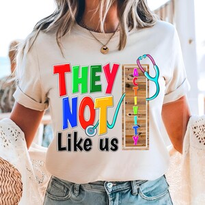 Puede incluir: Camiseta color crema con el texto "THEY NOT Like us" en letras coloridas y delineadas. Se incorpora un diseño de estetoscopio, con la palabra "ACTIVITY" alineada verticalmente. La camiseta tiene mangas cortas.