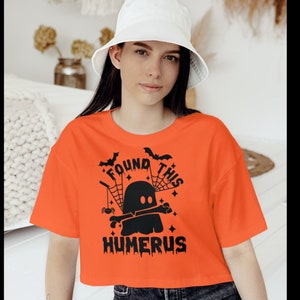 May include: An orange short-sleeved t-shirt featuring a black graphic print of a ghost holding a bone. The text reads I FOUND THIS HUMERUS, accompanied by illustrations of spider webs, a small spider, two bats, and several small stars.