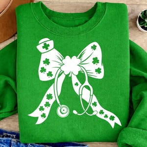 Op de afbeelding: Een groene sweatshirt met een wit strikdesign met een verpleegstershoed, klavers en een stethoscoop. De strik is het centrale element, met de stethoscoop eroverheen gedrapeerd. Klavers zijn verspreid over de strik.