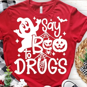 Puede incluir: Camiseta roja con un diseño blanco de Halloween. El diseño incluye un fantasma, calabazas, telarañas y la frase "Say Boo to Drugs". La camiseta está hecha de un material suave.