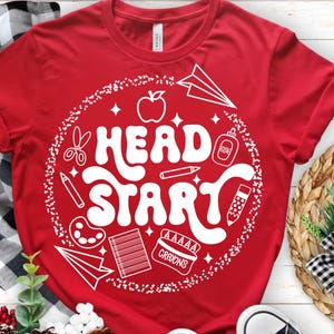 May include: Red t-shirt with a white graphic that says "Head Start" in a bubble font. The graphic includes school-themed illustrations like an apple, scissors, and crayons. The shirt is made of a soft material.