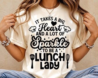 May include: A cream-colored t-shirt featuring black text that reads: IT TAKES A BIG HEART AND A LOT OF Sparkle TO BE A LUNCH LADY. The design includes two chef hats, a heart, small stars, a spatula, and a whisk graphic printed on the front.