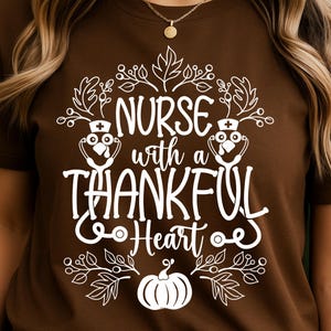 Può includere: T-shirt marrone con grafica bianca. La grafica dice "NURSE with a THANKFUL Heart" e include una zucca, foglie e un gufo con stetoscopio. Girocollo.