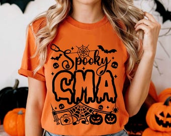 Op de afbeelding: Oranje t-shirt met de zwarte tekst "Spooky CMA", versierd met Halloween-motieven zoals vleermuizen, spinnenwebben, schedels en pompoenen. Het shirt heeft korte mouwen en een ronde hals, perfect voor Halloween.
