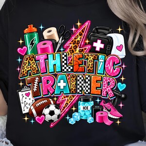 May include: A black t-shirt featuring a colourful graphic with the text ATHLETIC TRAINER. The design includes a leopard print lightning bolt, a water bottle, medical tape, scissors, a medical bag, a football, a soccer ball, ice, and a pink trainer.