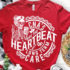 以下が含まれることがあります： 白いグラフィックが施された赤いTシャツ。デザインは、サングラスと看護師の帽子をかぶり、コーヒーカップを持ち、ピースサインをしている骸骨の看護師が特徴です。テキスト:「CNA's are the HEARTBEAT of LONG TERM CARE。」