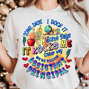 May include: White t-shirt with a colourful graphic design that says "Some days I rock it, some days it rocks me, either way we're rockin' assistant principal".
