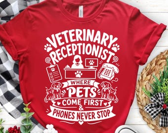 May include: A red t-shirt with a white graphic that reads "Veterinary Receptionist" with paw prints, a desk, a phone, and the text "Where Pets Come First & Phones Never Stop". The shirt features a crew neck and short sleeves.