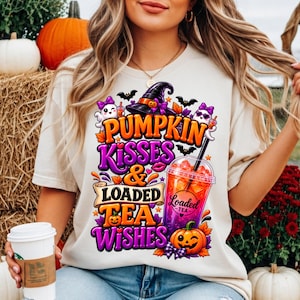 May include: A cream-coloured t-shirt featuring a colourful graphic with the text PUMPKIN KISSES & LOADED TEA WISHES. The design includes a witch hat, ghosts, bats, autumn leaves, a jack-o'-lantern, and a plastic cup filled with a vibrant drink.