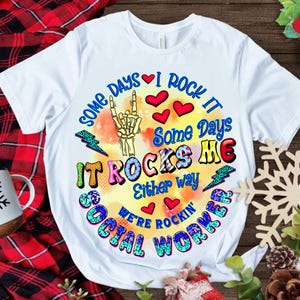 Peut inclure: T-shirt blanc avec un motif graphique coloré qui dit "Some days I rock it, some days it rocks me, either way we're rockin' social worker".