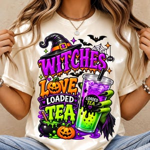 May include: A cream-colored t-shirt featuring a graphic print with the text "WITCHES LOVE LOADED TEA". The design includes a witch hat, two jack-o'-lanterns, a black cauldron, bats, a spider web, and a green hand holding a cup of loaded tea.