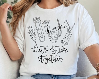 Könnte beinhalten: Weißes T-Shirt mit einer schwarzen Strichzeichnung von medizinischen Instrumenten mit lächelnden Gesichtern, darunter eine Spritze und Reagenzgläser. Der Schriftzug "Let's Stick Together" steht darunter.