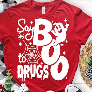 Puede incluir: Una camiseta roja de manga corta con un estampado gráfico blanco. El diseño incluye una telaraña, un pequeño fantasma y el texto "Say BOO to DRUGS" en una mezcla de fuentes negritas, estilizadas y goteantes, acentuado por pequeñas estrellas.