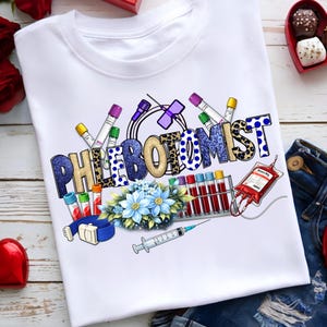 Puede incluir: Camiseta blanca con la palabra "PHLEBOTOMIST" en un diseño colorido. El diseño incluye equipos médicos como tubos de ensayo, una bolsa de sangre y una jeringa, junto con flores azules.