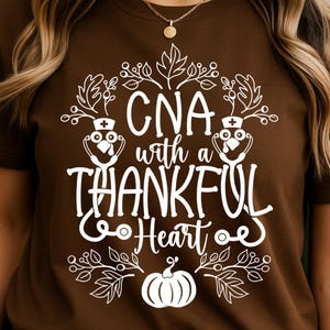 May include: A brown t-shirt featuring a white graphic that says "CNA with a THANKFUL Heart." The design includes a stethoscope, a pumpkin, and leaves. The shirt has a crew neck.