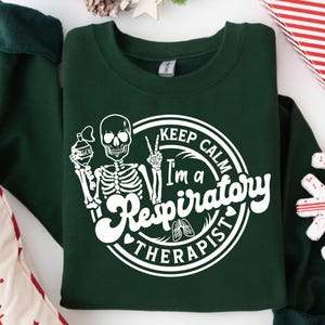 May include: Dark green sweatshirt with a white graphic of a skeleton holding an oxygen tank and making a peace sign. The text reads "Keep Calm I'm a Respiratory Therapist."
