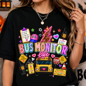 May include: A black t-shirt featuring a colorful graphic design with a yellow school bus, a leopard print lightning bolt, a clipboard, a stop sign, a traffic cone, and two-way radios. The text reads BUS MONITOR life, along with various phrases.