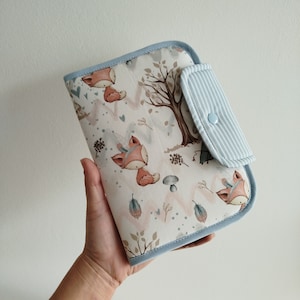 Könnte beinhalten: Eine hellblaue Wickelunterlage mit weißem Hintergrund und einem Muster aus Füchsen, Bäumen und Blättern. Die Unterlage hat eine kleine Tasche aus blau-weiß gestreiftem Stoff mit einem blauen Knopf.
