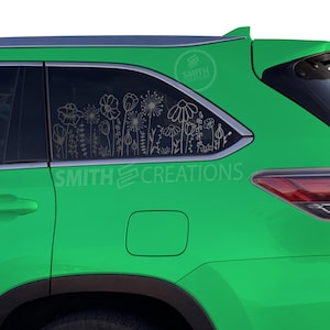 May include: A green SUV with a floral design on the back window. The design is a silhouette of flowers and leaves. The car has a black rear spoiler and a black gas cap.