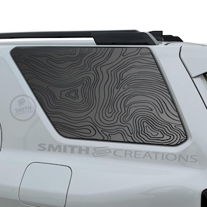 May include: White SUV with a black topographical map design on the rear window. The text "SMITH CREATIONS" is visible on the vehicle. A black roof rack is also present.