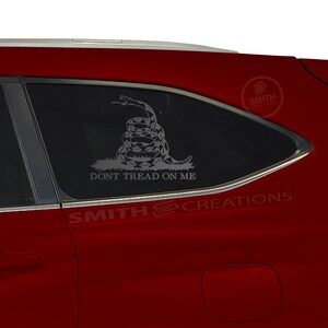 May include: A red car with a window decal of a snake coiled around a circle with the text "Don't Tread On Me". The decal is in gray and the text is in white. The car has a black window trim and the text "Smith Creations" is visible on the bottom of the image.