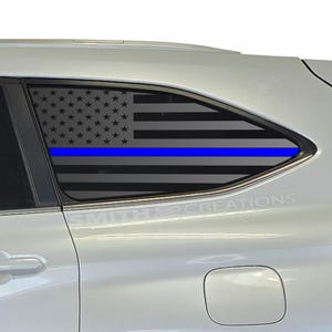 May include: A car window decal featuring the American flag with a blue line, representing support for law enforcement. The stars and stripes are in shades of gray, with a vibrant blue stripe. The decal is applied to a car window.