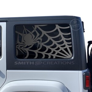 May include: A black and gray spider web and spider decal applied to the rear window of a white Jeep. The decal is a silhouette design. The text "SMITH CREATIONS" is below the window.