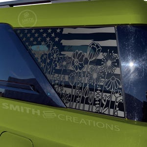 May include: A green vehicle with a window decal featuring an American flag design with a floral overlay. The decal includes the text "SMITH CREATIONS". The design is in black and white, with the flag's stars and stripes visible.