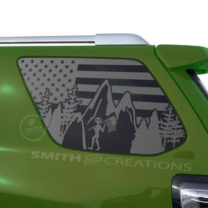 May include: A green vehicle with a window decal featuring an American flag design, mountains, trees, and an alien holding a flag. The decal is gray and the text "SMITH CREATIONS" is visible.