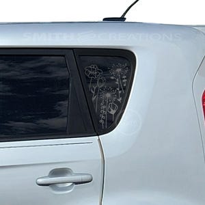 May include: A silver car with a floral decal on the rear window. The decal features various flower designs, including poppies and dandelions. The car has a black antenna and a red taillight.