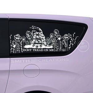 May include: White vinyl car decal featuring a coiled rattlesnake and floral elements, with the text "DON'T TREAD ON ME." The decal is applied to a car window. The brand name "SMITH CREATIONS" is also visible.