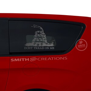 May include: A red car with a black window sticker featuring a coiled snake with the text "Don't Tread On Me". The sticker is on the back passenger side window. The sticker also includes the text "Smith Creations".
