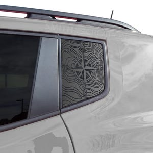 May include: A car window decal featuring a black compass rose design with topographic lines. The decal is applied to the rear side window of a light-colored vehicle. The design includes the letters N, E, W, and S, indicating the cardinal directions.