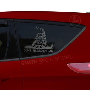 May include: A red car with a white vinyl sticker on the back window. The sticker features a coiled snake with the words "Don't Tread On Me".