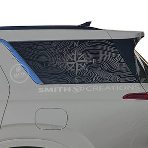 May include: A white SUV with a custom window decal featuring a topographic map design and a compass rose. The decal is black and white, with the text "SMITH CREATIONS" visible on the vehicle.