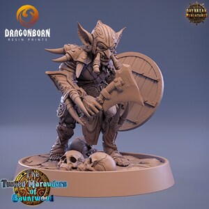 May include: A detailed miniature of a goblin warrior, painted in a brown colour scheme. The goblin is holding an axe and a shield, and is standing on a base with skulls. The miniature is part of the "The Tusked Marauders of Gauntwood" series.