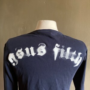 May include: A navy blue long-sleeve shirt with a white spray-painted graphic that says "Jesus Filth" on the back.