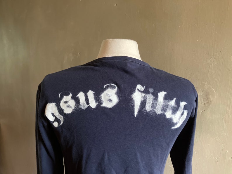 May include: A navy blue long-sleeve shirt with a white spray-painted graphic that says "Jesus Filth" on the back.