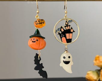 May include: Asymmetrical dangle earrings featuring Halloween motifs. One earring has a small orange pumpkin, a larger pumpkin wearing a green hat, and a black bat. The other features a gold circular frame with a black haunted house and a white ghost.