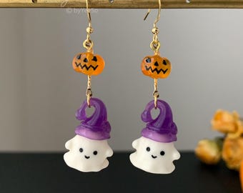 May include: A pair of drop earrings featuring a small orange jack-o'-lantern bead topped with a white ghost wearing a purple hat. The charms are suspended from gold-toned metal hooks. The text "yeeso" and "byYeeSo" is visible at the top.